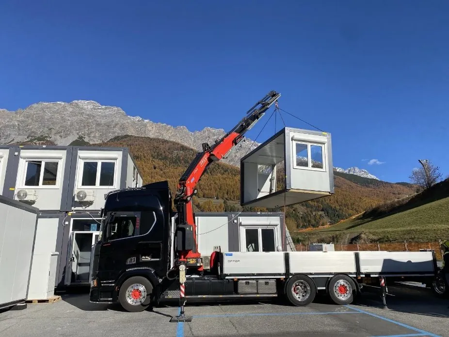 Camion con gru che posa un modulo prefabbricato accanto ad altri moduli, montagne sullo sfondo