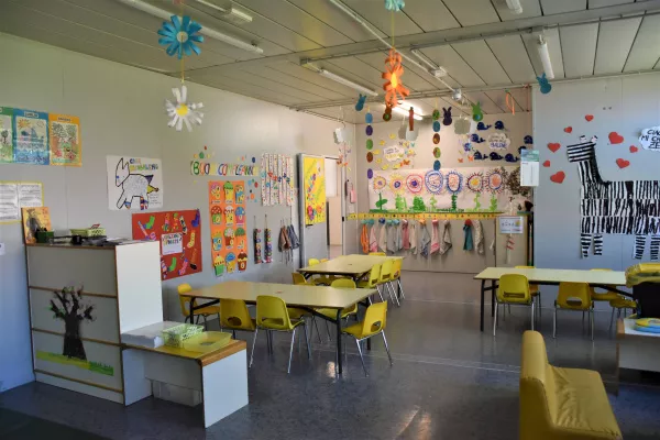 Sezione di scuola dell'infanzia con banchi sedie gialle e disegni appesi